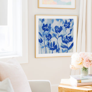 a cozy living room with a large blue floral painting hanging on the wall, a white chair, a table with a vase of pink flowers, and a stack of books.