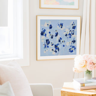 a cozy living room with a white chair, a framed painting of blue flowers on the wall, and a vase of pink flowers on a wooden table.