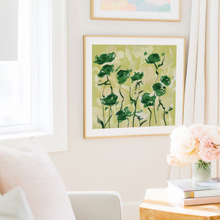 a cozy living room with a framed painting of green flowers hanging on the wall, a vase of pink flowers on a table, and a bookshelf filled with books.