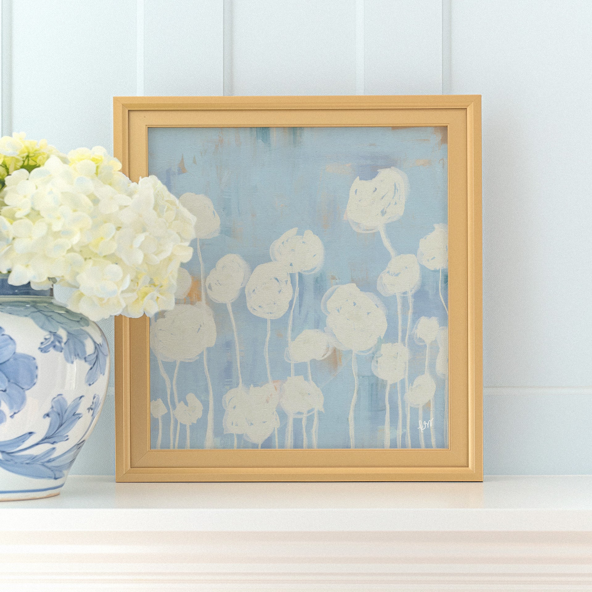a framed artwork of white flowers on a white shelf, accompanied by a blue and white vase filled with white hydrangeas.