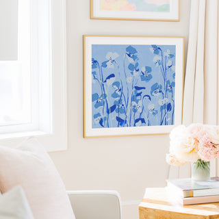 a cozy living room with a white sofa, a framed artwork of blue flowers on the wall, and a vase of pink flowers on a wooden table.