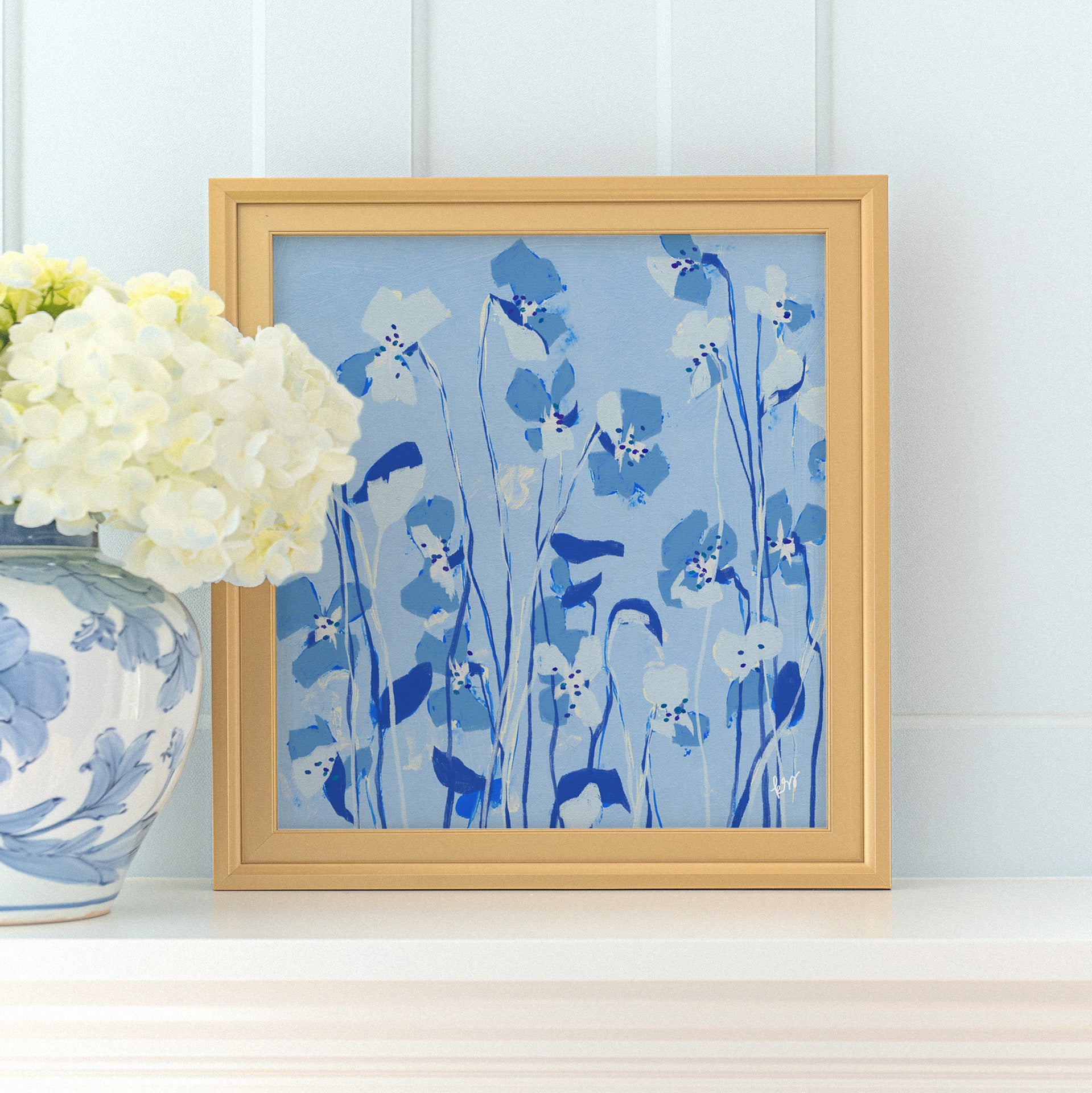 a framed painting of blue flowers on a white shelf, with a vase of white hydrangeas placed next to it.