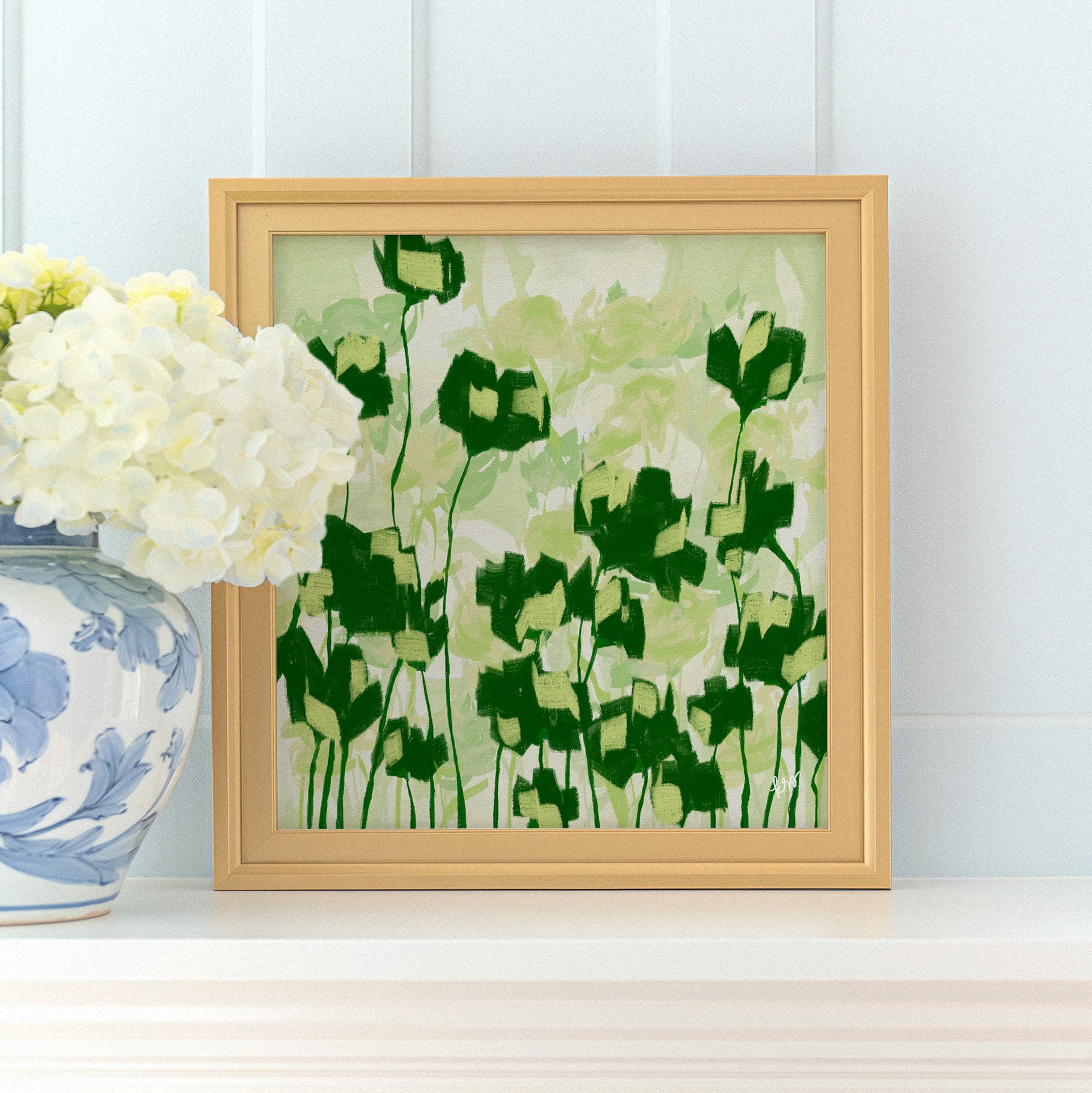 a framed painting of green and white flowers on a white shelf, with a vase of white hydrangeas placed next to it.