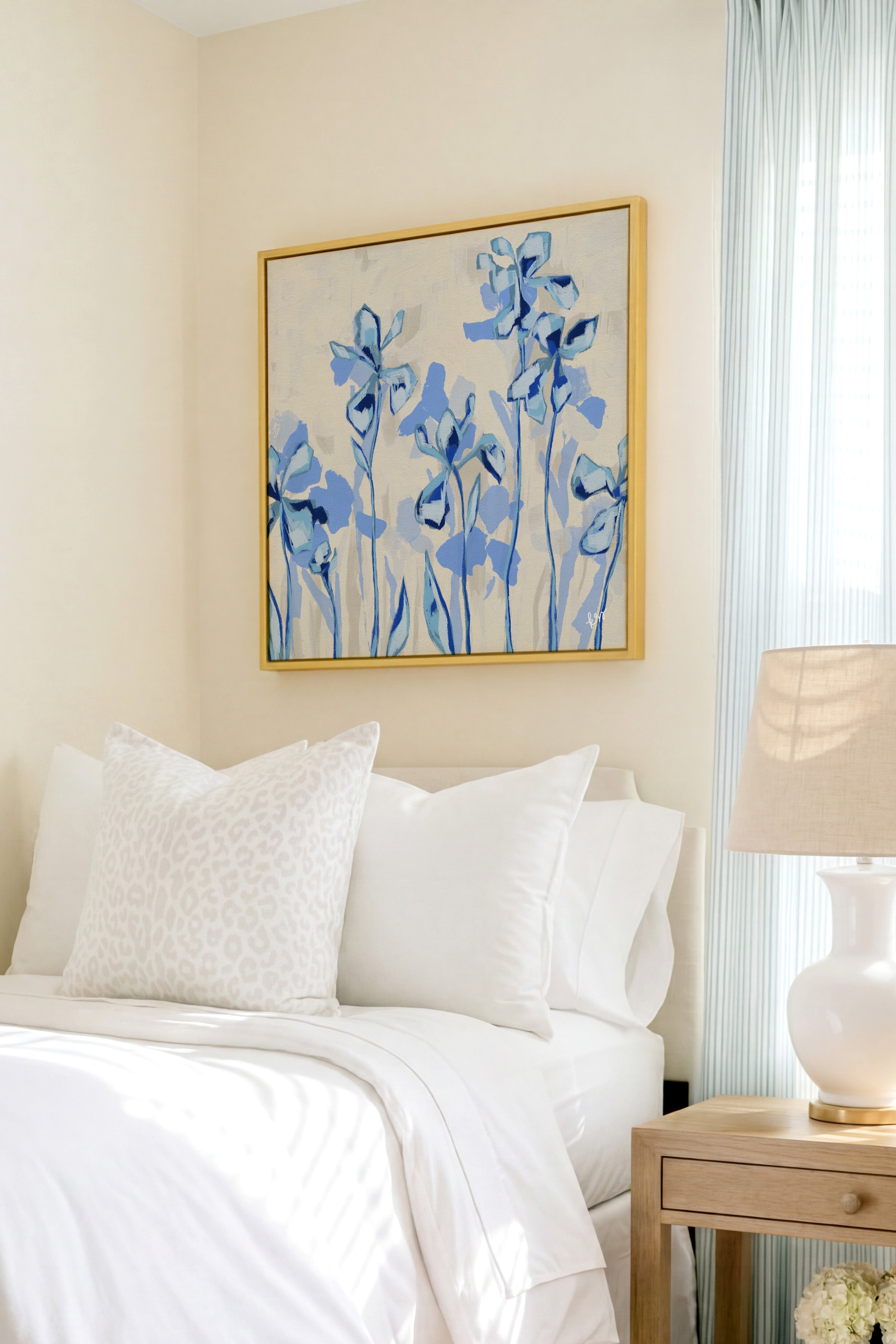 a serene bedroom scene with a bed adorned with white linens and a framed painting of blue flowers hanging above it. The painting adds a touch of color and life to the otherwise neutral-toned room.