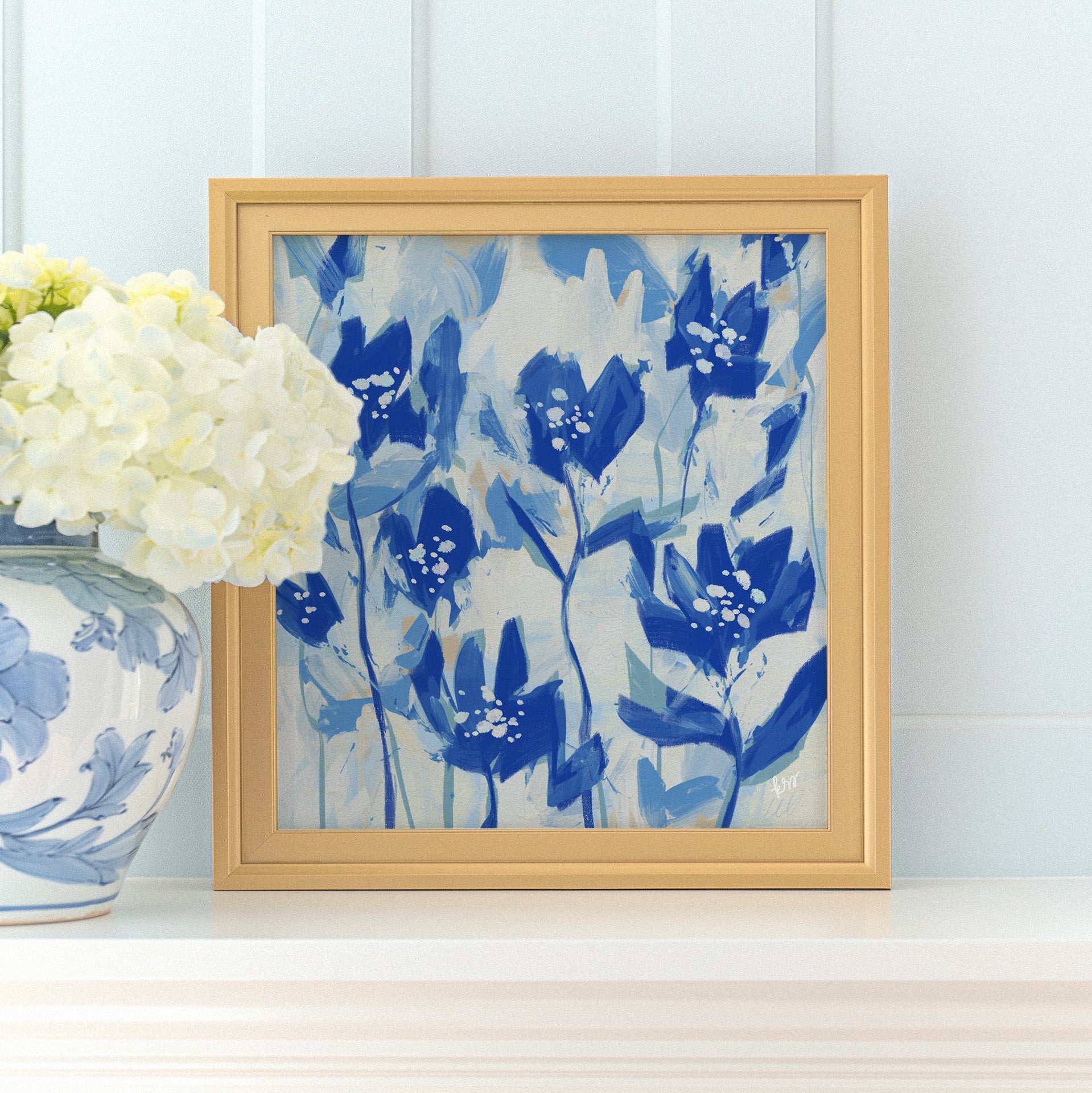 a framed painting of blue flowers on a white shelf, with a vase of white hydrangeas placed next to it.