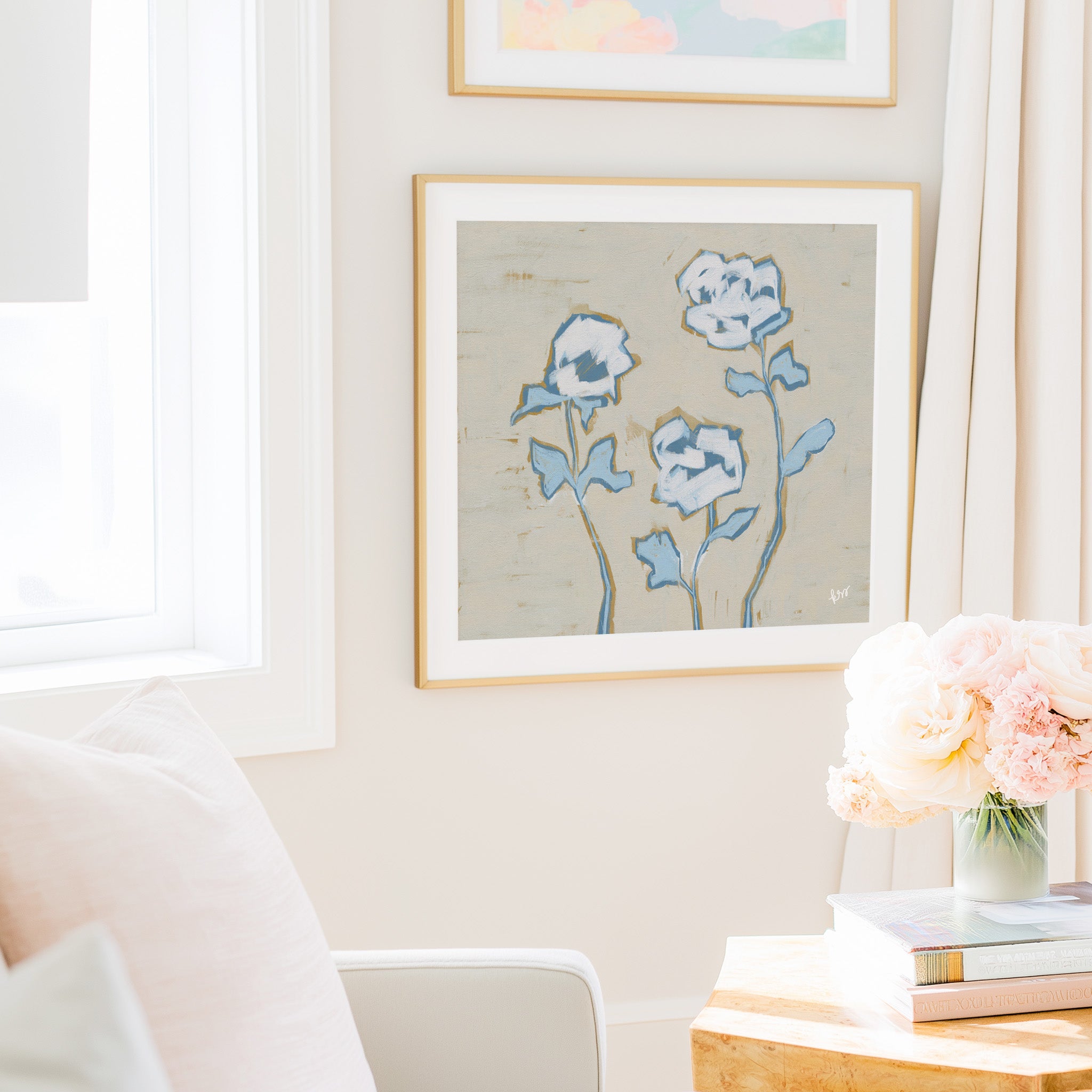 a cozy living room with a framed painting of blue flowers hanging on the wall, a vase of pink flowers on a wooden table, and a stack of books on the table.