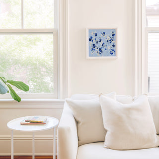a cozy living room with a white sofa, a small white side table, and a framed artwork on the wall. The room is bright and airy, with natural light coming in through a window.