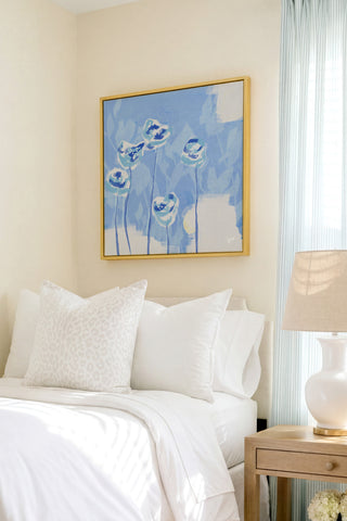 a serene bedroom scene with a bed adorned with white pillows and a white comforter, complemented by a framed painting of blue flowers hanging above it. The room is further enhanced by a wooden nightstand and a lamp, creating a cozy and inviting atmosphere.