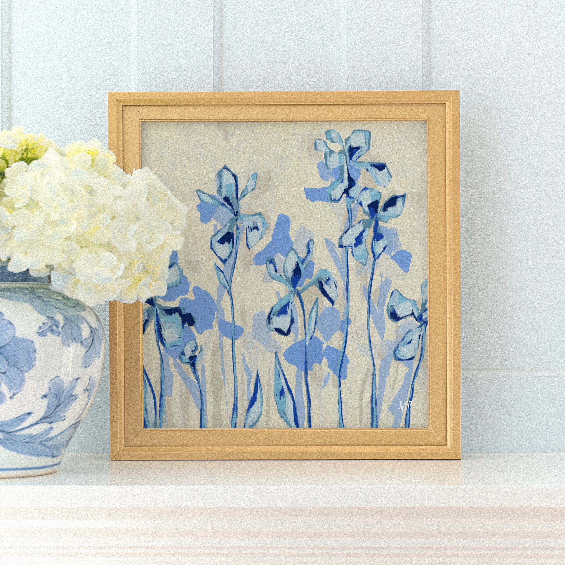 a framed painting of blue flowers on a white shelf, with a vase of white hydrangeas placed next to it.