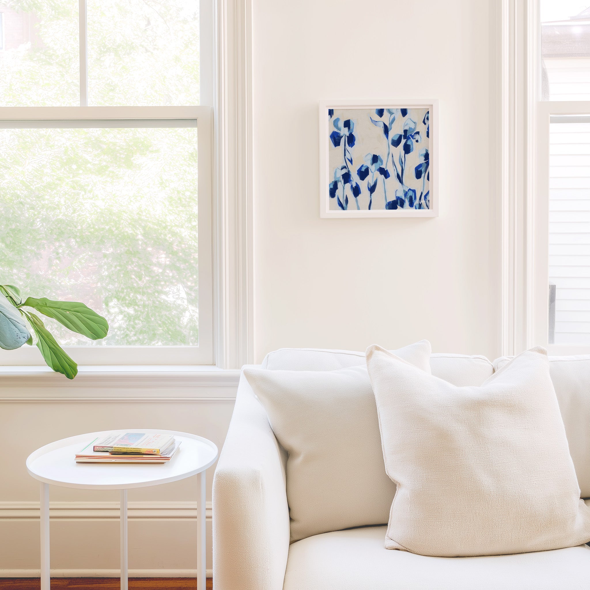a cozy living room with a white sofa, a small round table, and a framed artwork featuring blue flowers on the wall.