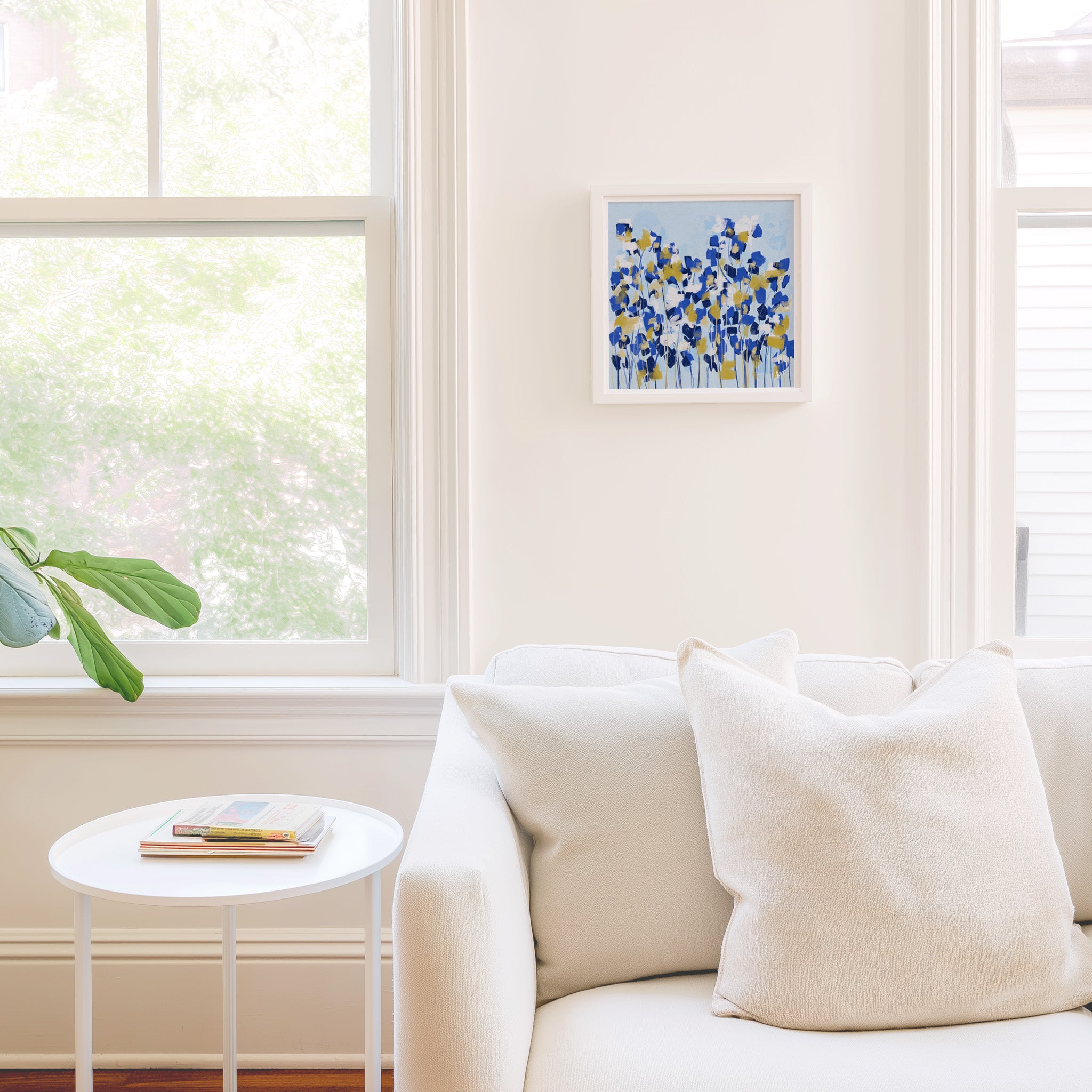 a cozy living room with a white sofa, a small round table, and a framed artwork of blue and yellow flowers on the wall.