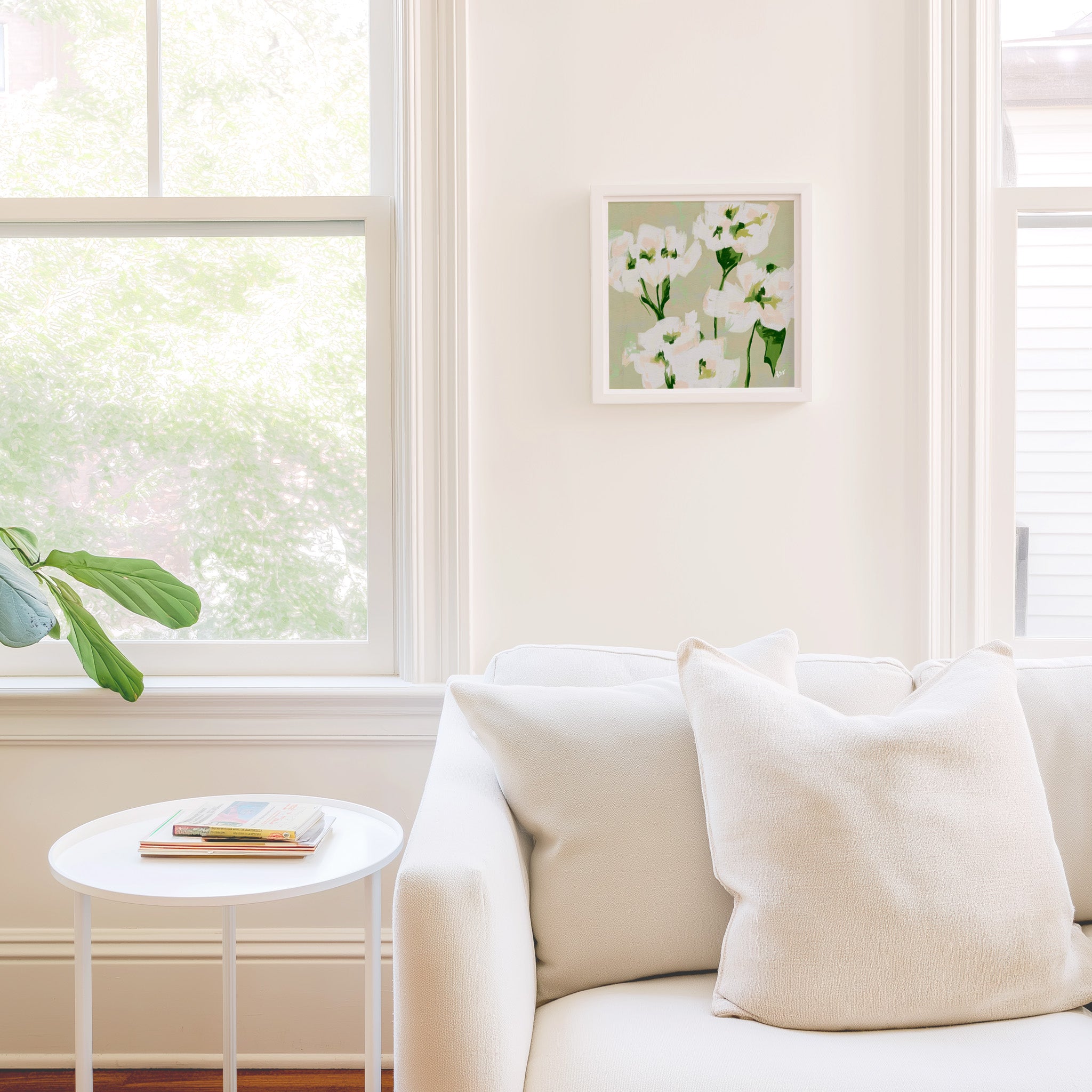 a cozy living room with a white sofa, a small white side table, and a framed picture of white flowers hanging on the wall.