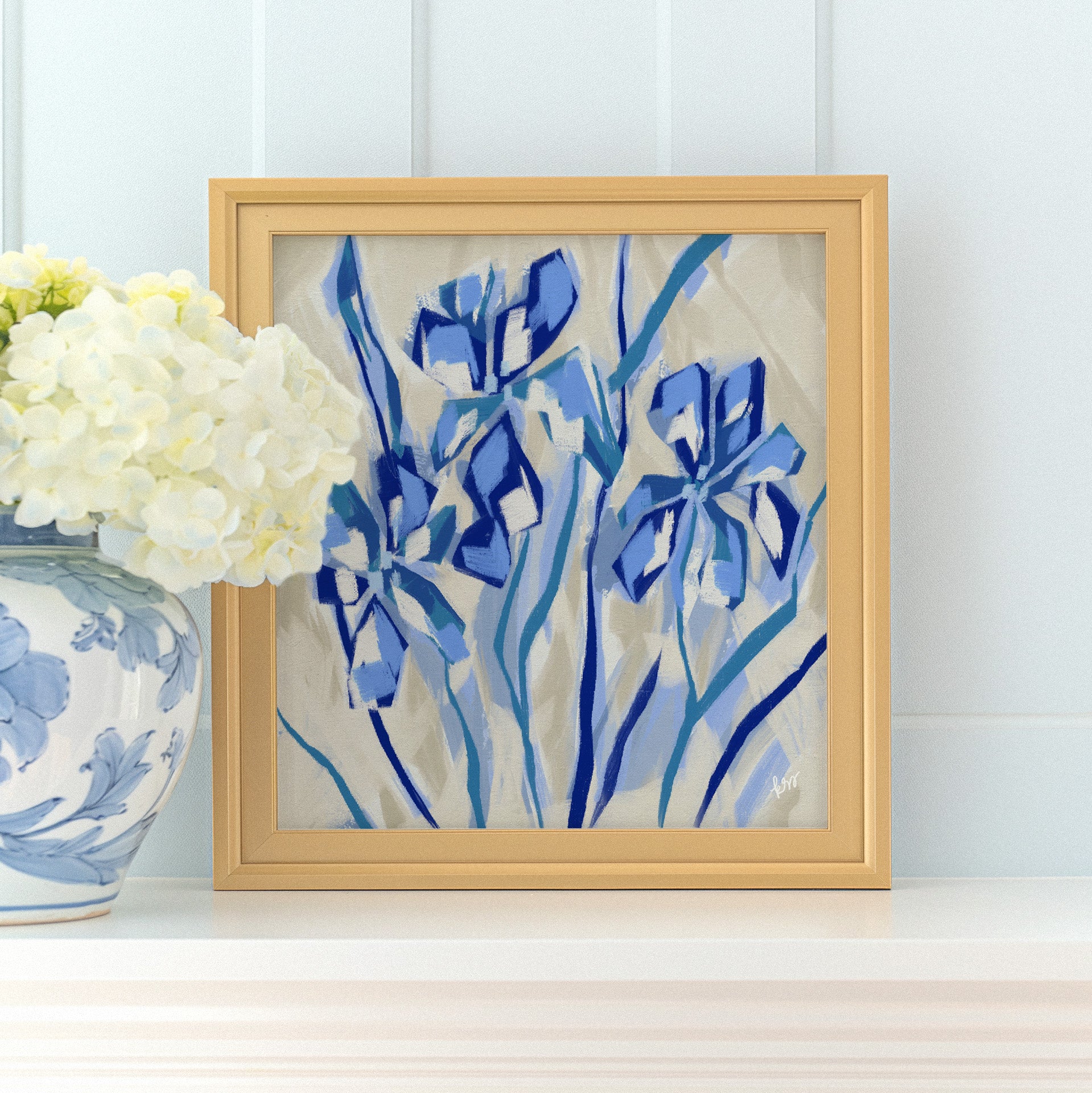 a framed painting of blue flowers on a white shelf, with a vase of white hydrangeas placed next to it.