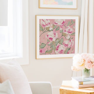 a cozy living room with a large framed floral print hanging on the wall, a vase of pink flowers on a table, and a bookshelf filled with books.