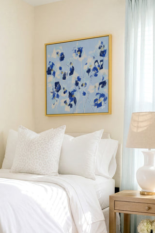 a serene bedroom scene with a bed adorned with white pillows and a blue floral painting hanging above it. The painting adds a touch of color and life to the otherwise neutral-toned room.
