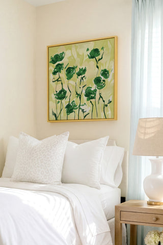 a serene bedroom scene with a bed adorned with white pillows and a white comforter, complemented by a framed artwork featuring green flowers hanging above it.