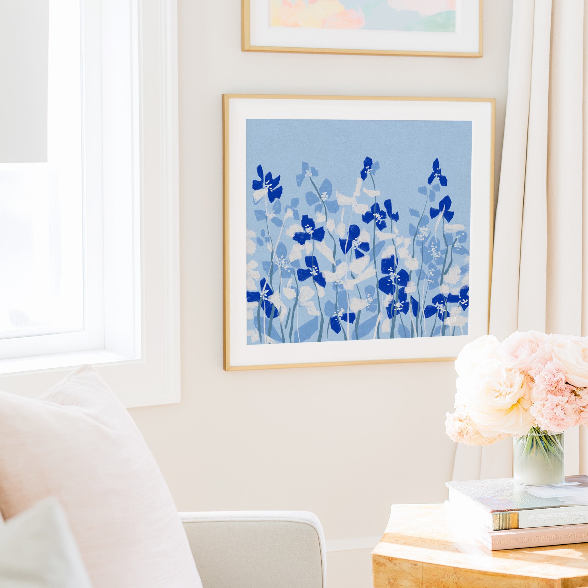 a cozy living room with a framed artwork of blue flowers hanging on the wall, a vase of pink flowers on a table, and a bookshelf filled with books.