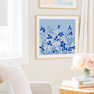 a cozy living room with a framed artwork of blue flowers hanging on the wall, a vase of pink flowers on a table, and a bookshelf filled with books.