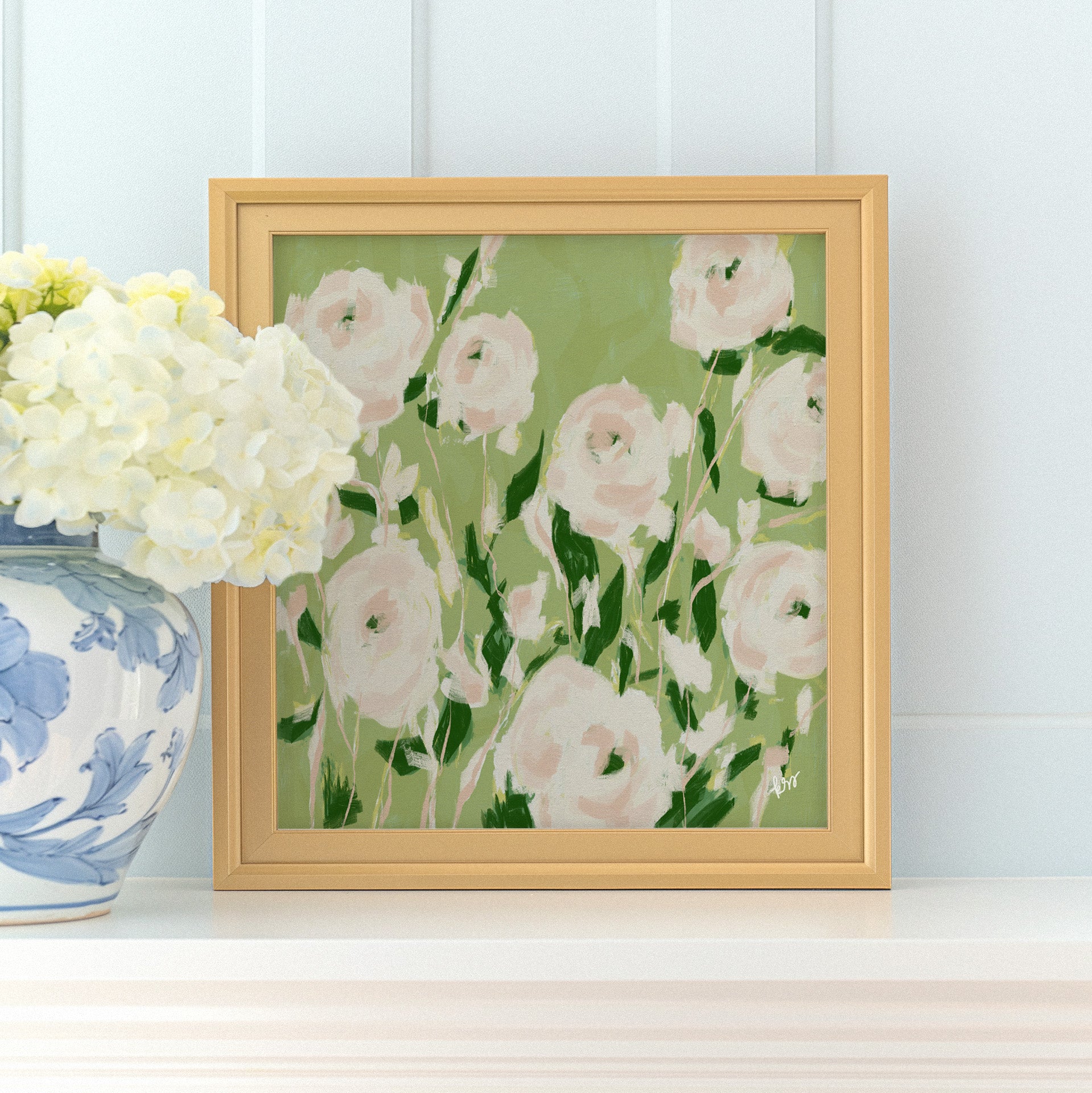 a framed painting of white and green flowers on a white shelf, with a blue and white vase of white hydrangeas placed next to it.