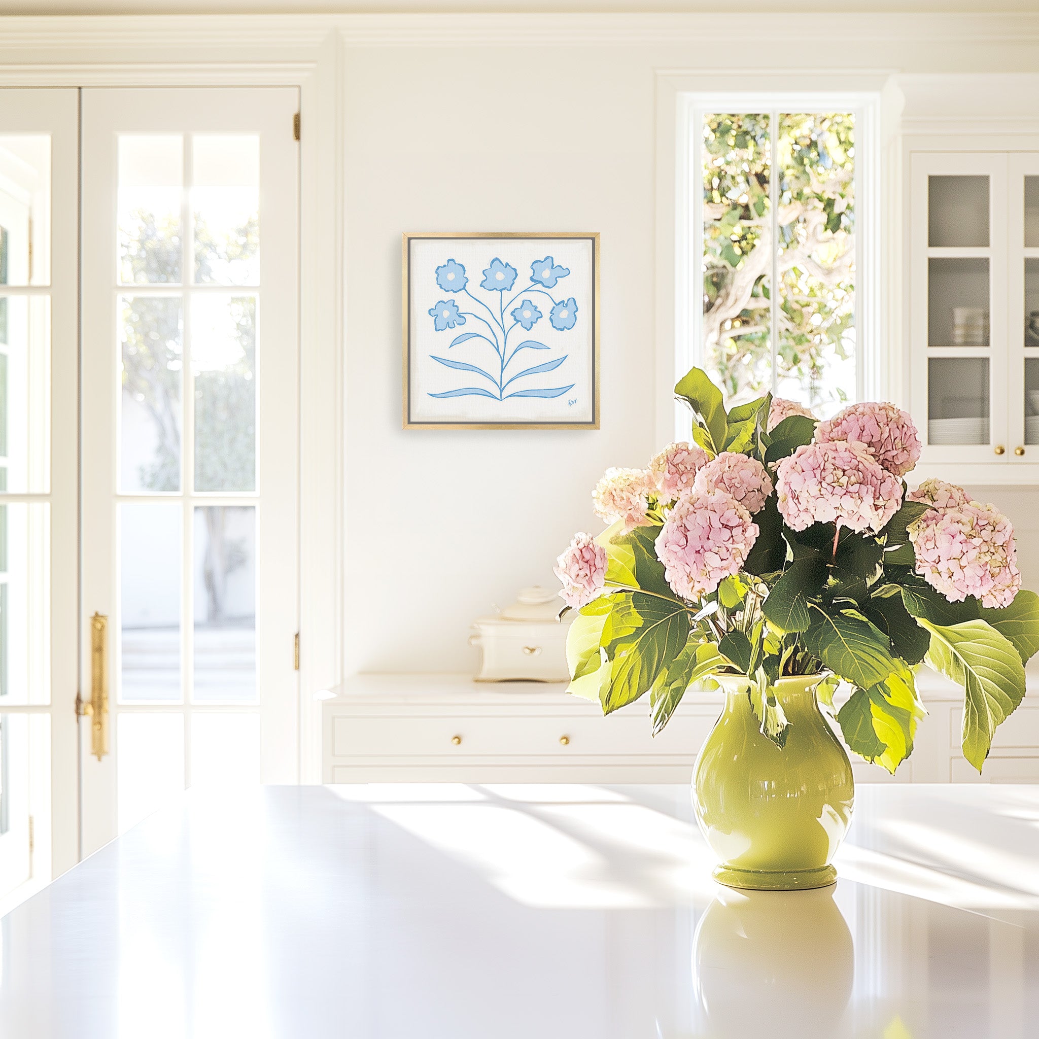 a white kitchen with a vase of pink hydrangeas on the counter, a framed blue floral artwork on the wall, and a window letting in natural light.