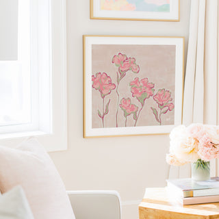 a cozy living room with a framed floral artwork hanging on the wall, a vase of pink flowers on a table, and a bookshelf filled with books.
