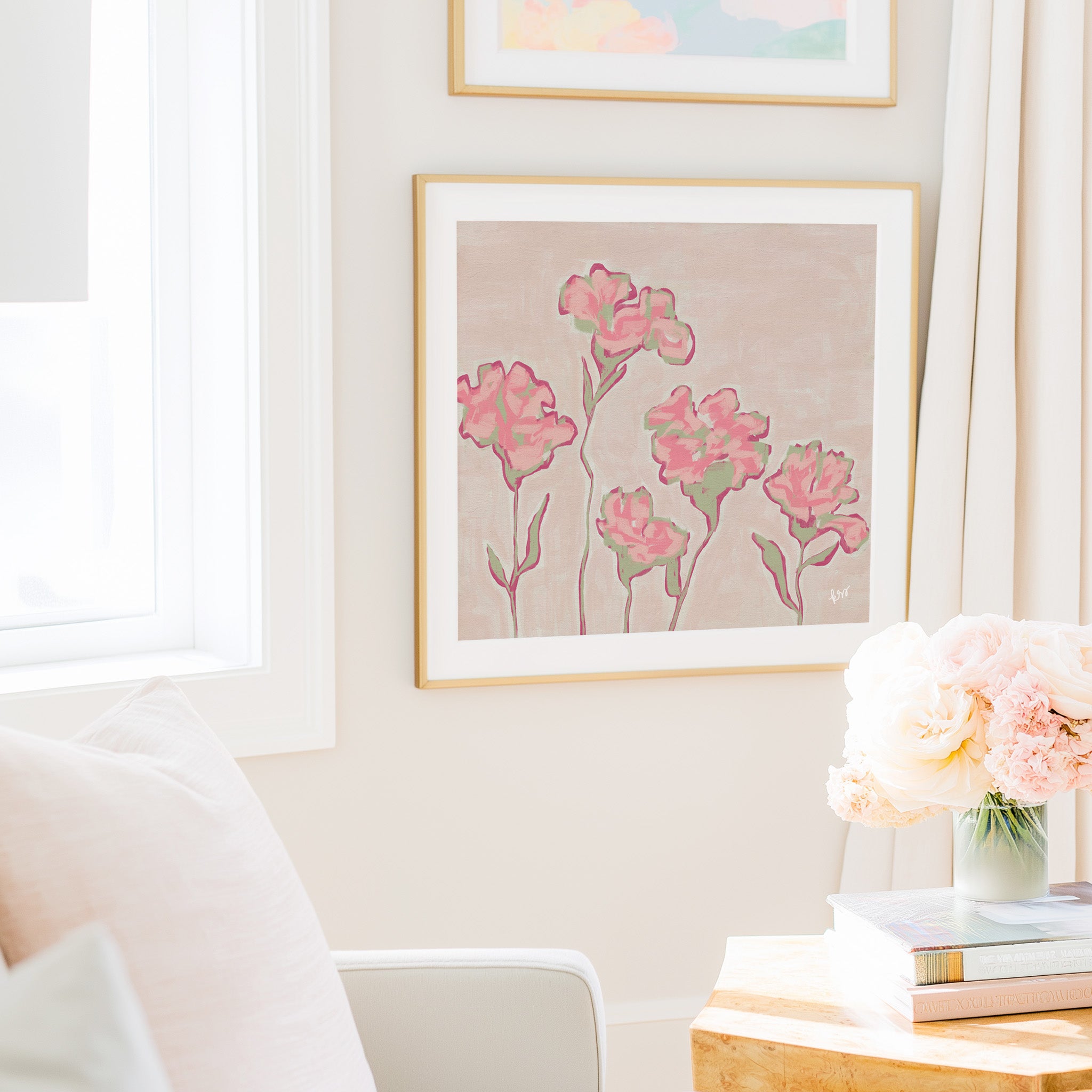 a cozy living room with a framed floral artwork hanging on the wall, a vase of pink flowers on a table, and a bookshelf filled with books.