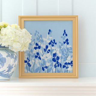 a framed artwork of blue flowers on a white shelf, with a vase of white hydrangeas placed next to it.