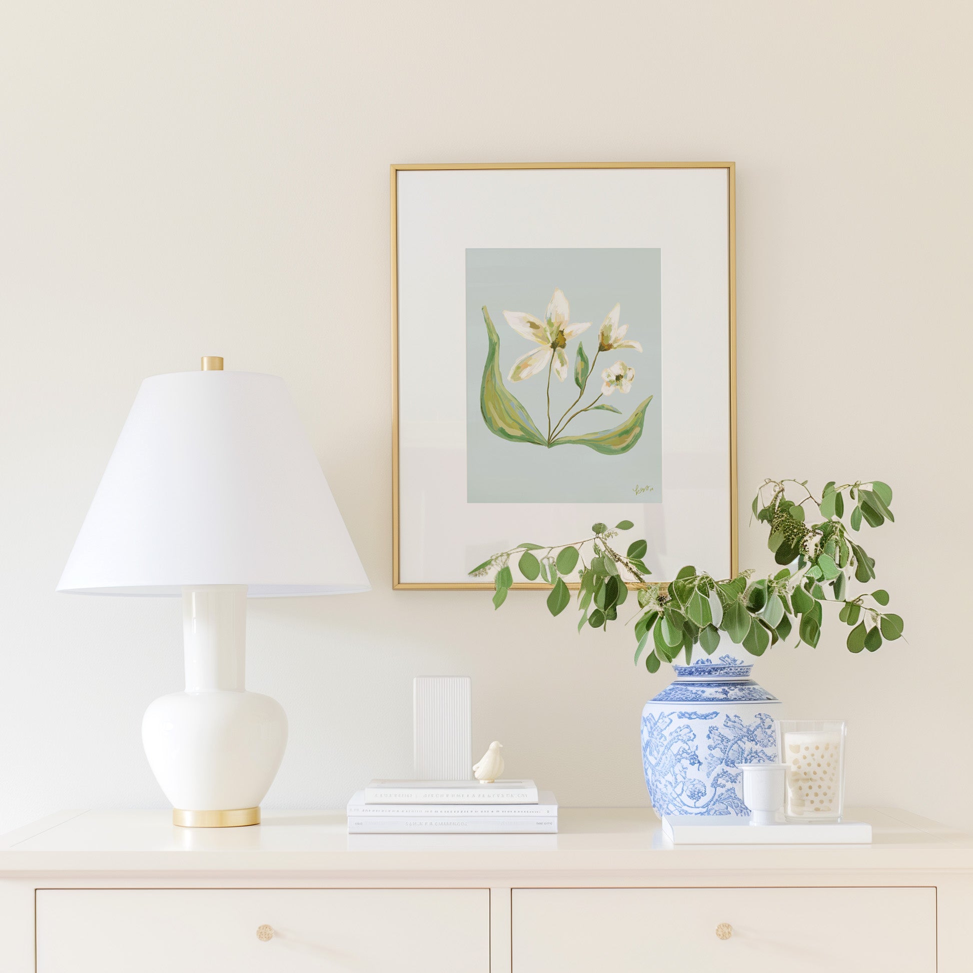 a white dresser with a framed picture of white flowers, a white lamp, a blue and white vase with greenery, and some books and a small bird figurine on top of the dresser.