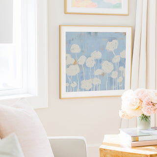 a cozy living room with a white sofa, a framed artwork of white flowers on the wall, and a vase of pink flowers on a wooden table.