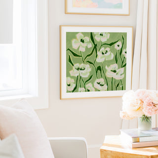 a cozy living room with a large framed painting of white flowers hanging on the wall, a white sofa, a wooden side table with a vase of pink flowers, and a stack of books on the table.