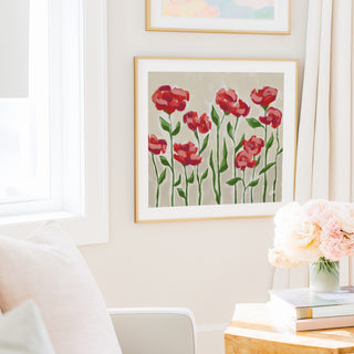 a cozy living room with a framed painting of red flowers hanging on the wall, a white chair, a table with a vase of pink flowers, and a stack of books.