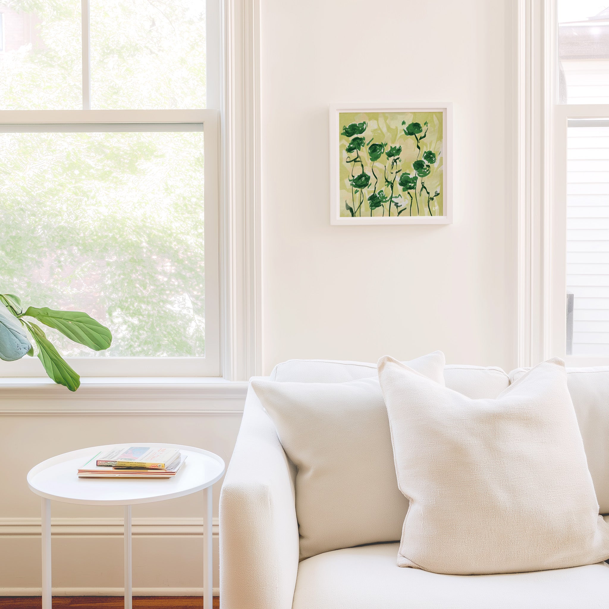 a cozy living room with a white sofa, a small round table, and a framed picture of green flowers on the wall.