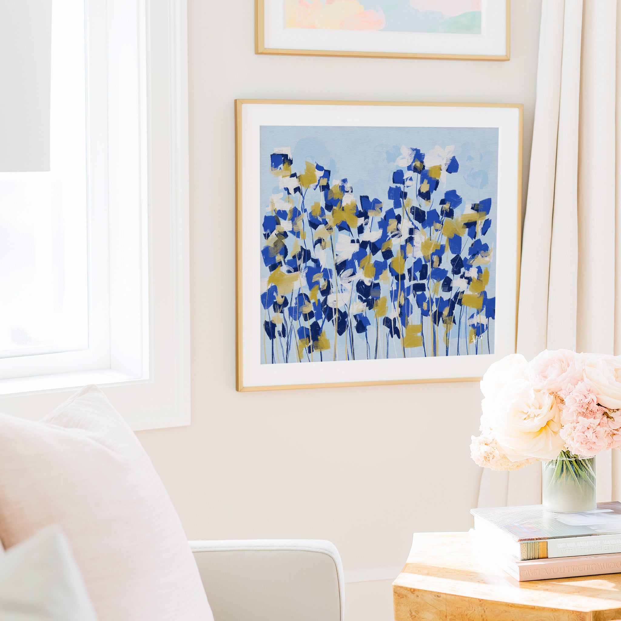 a cozy living room with a white sofa, a framed painting of blue and yellow flowers hanging on the wall, and a vase of pink flowers on a wooden table.