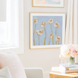 a cozy living room with a framed painting of white flowers hanging on the wall, a vase of pink flowers on a table, and a bookshelf filled with books.