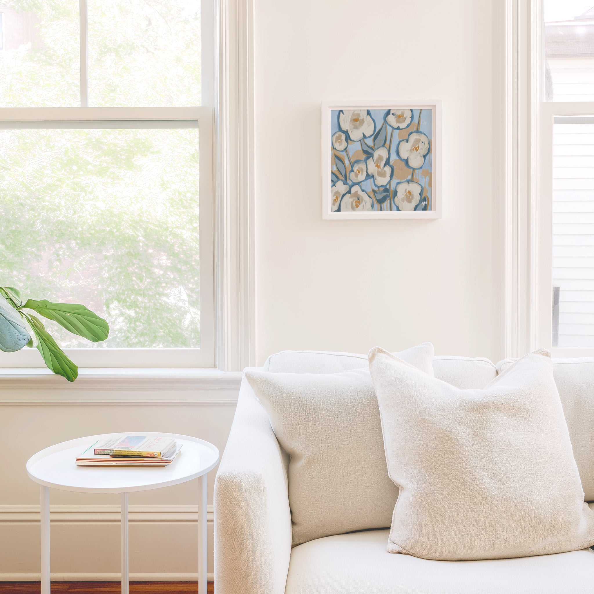 a cozy living room with a white sofa, a small round table, and a framed artwork on the wall. The room is bright and airy, with natural light coming in through a window.