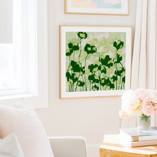 a cozy living room with a white sofa, a framed artwork of green flowers on the wall, and a vase of pink flowers on a wooden table.