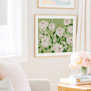 a cozy living room with a large framed painting of pink flowers hanging on the wall, a white chair, a wooden side table with a vase of pink flowers, and a stack of books on the table.