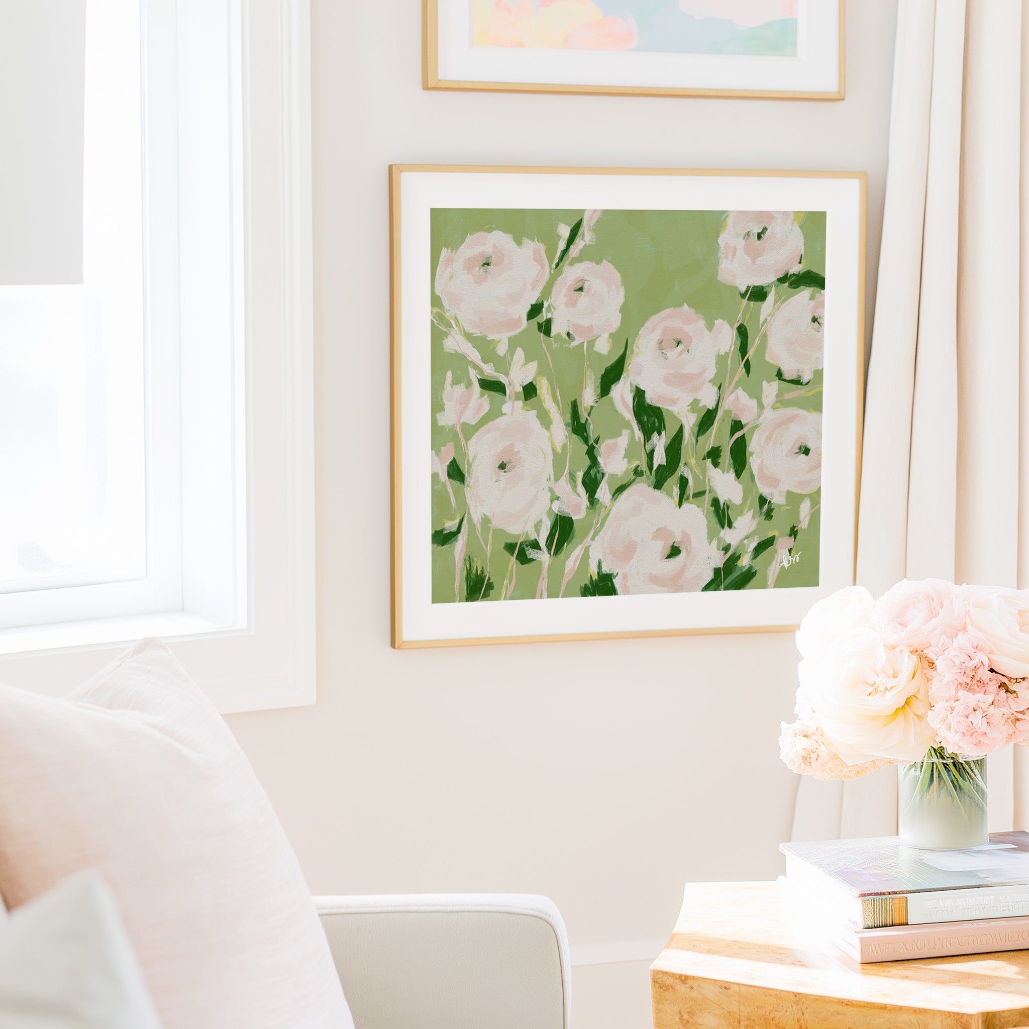 a cozy living room with a large framed painting of pink flowers hanging on the wall, a white chair, a wooden side table with a vase of pink flowers, and a stack of books on the table.