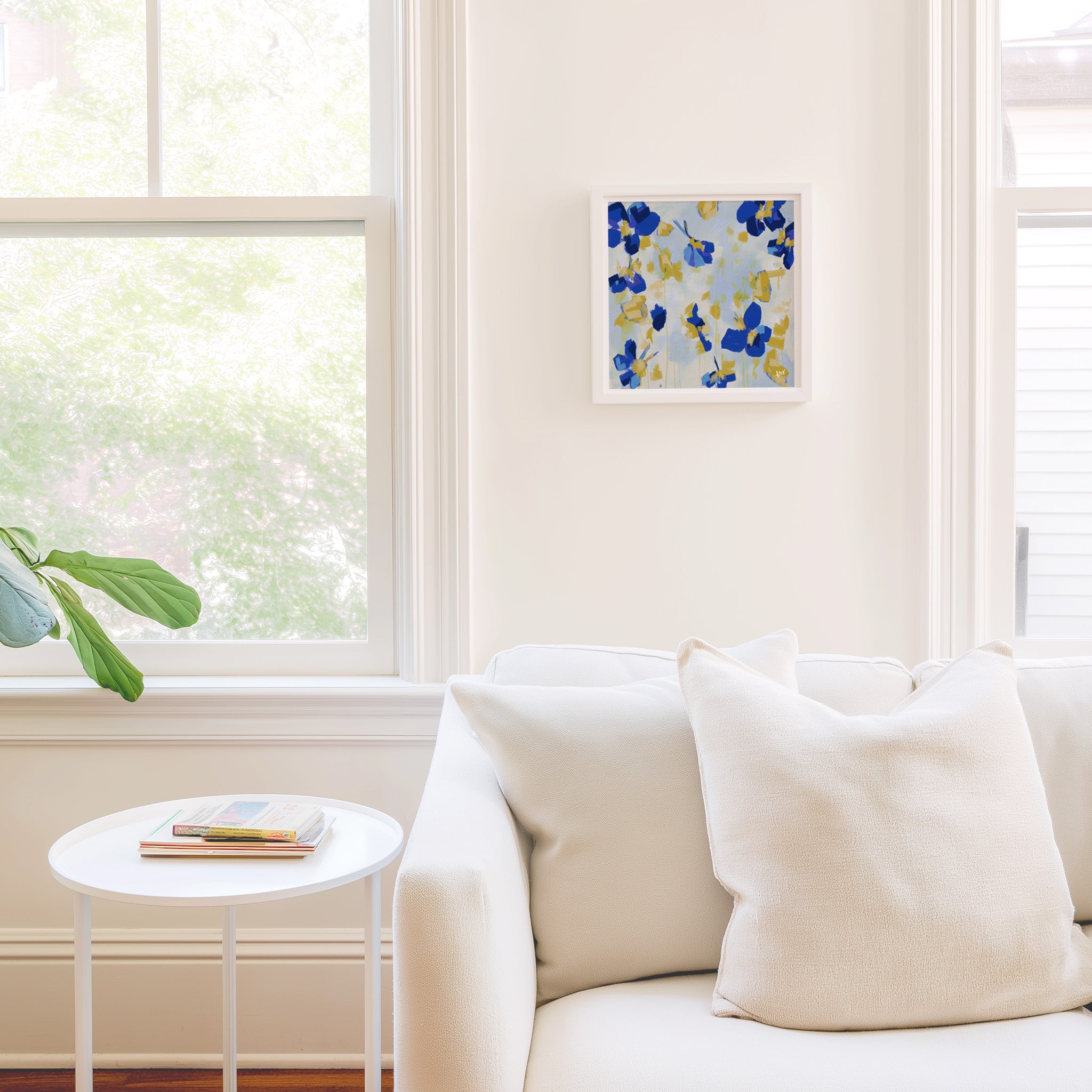 a cozy living room with a white sofa, a small round table, and a framed artwork on the wall. The room is bright and airy, with natural light coming in through a window.