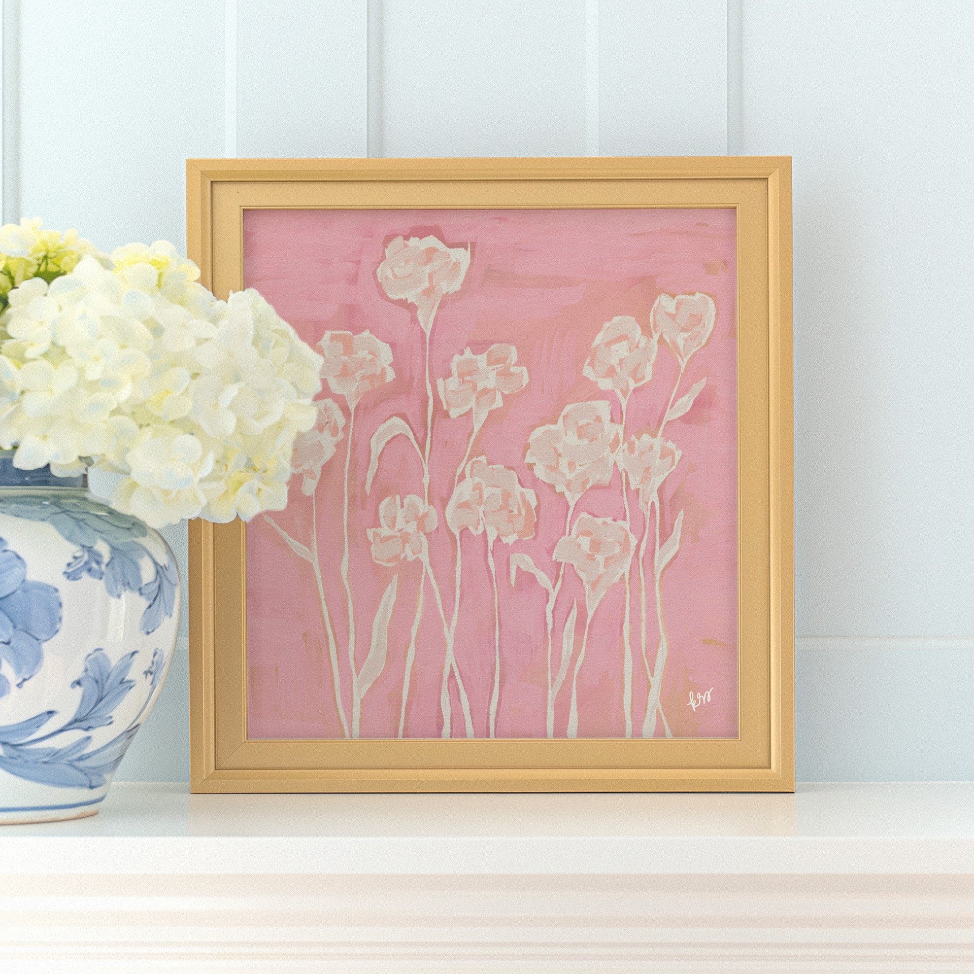 a framed painting of white flowers on a pink background, placed on a white shelf next to a blue and white vase filled with white hydrangeas.