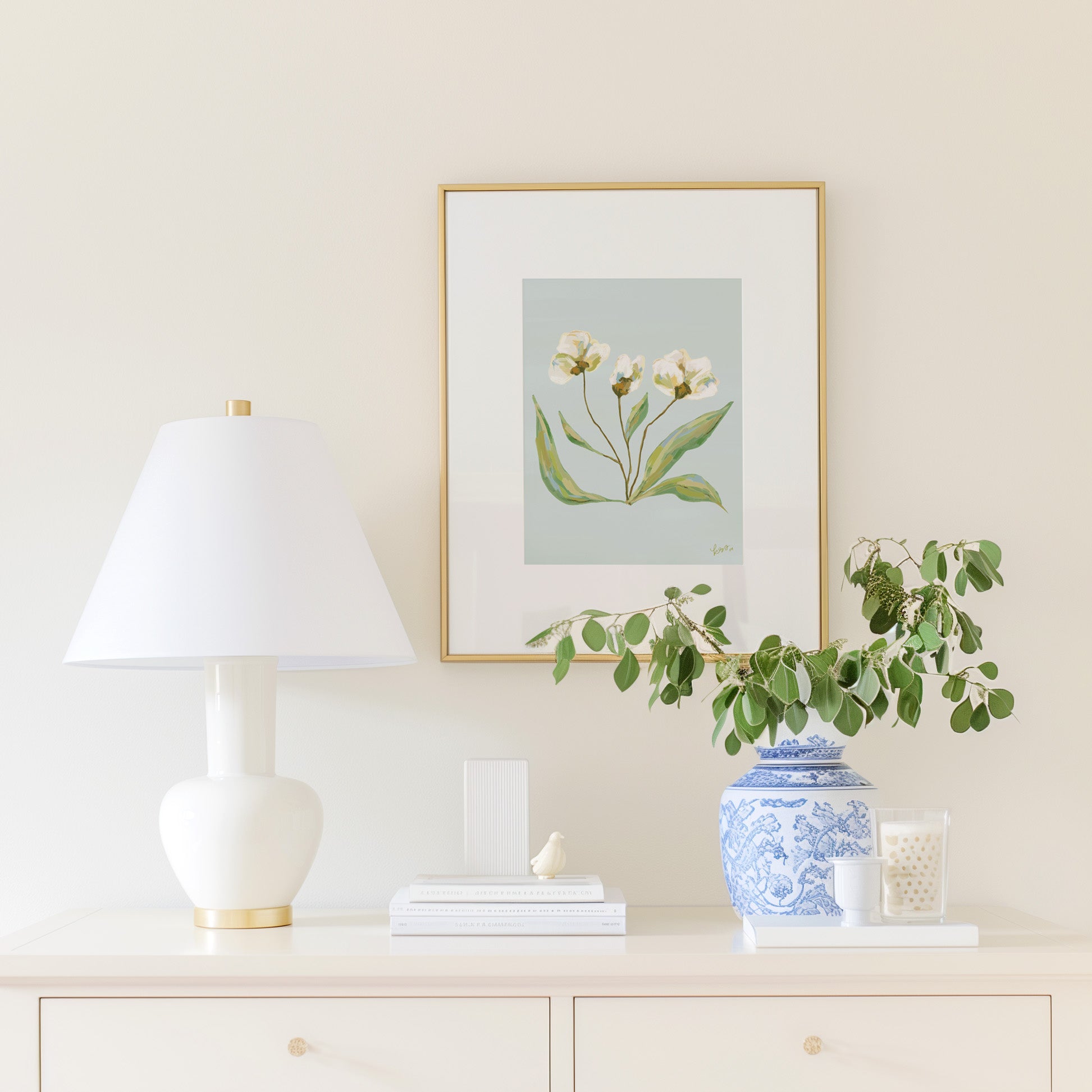 a white dresser with a framed picture of white flowers, a white lamp, a blue and white vase with greenery, and some books and a small bird figurine on top of the dresser.