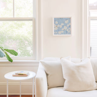 a cozy living room with a white sofa, a small white side table, and a framed artwork on the wall. The room is bright and airy, with natural light coming in through a window.