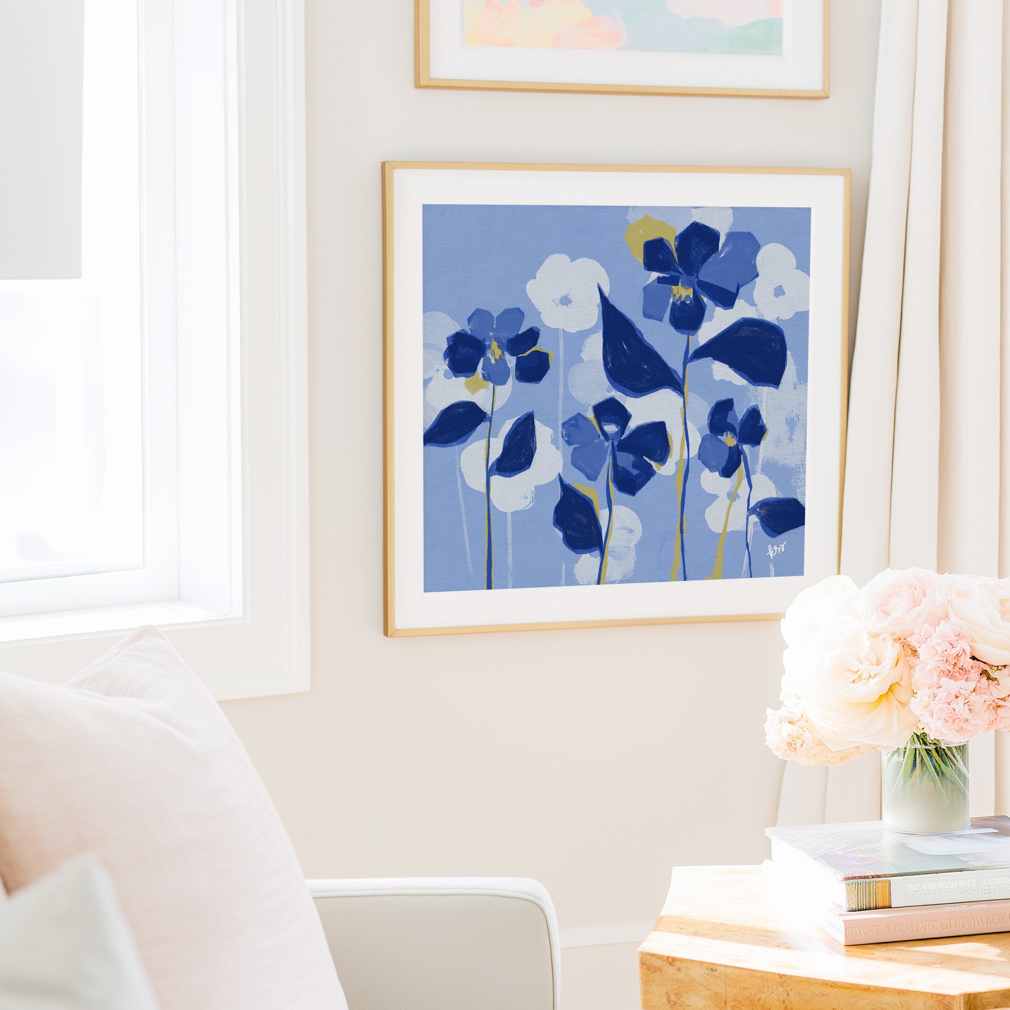 a cozy living room with a framed artwork of blue flowers hanging on the wall, a vase of pink flowers on a table, and a bookshelf filled with books.