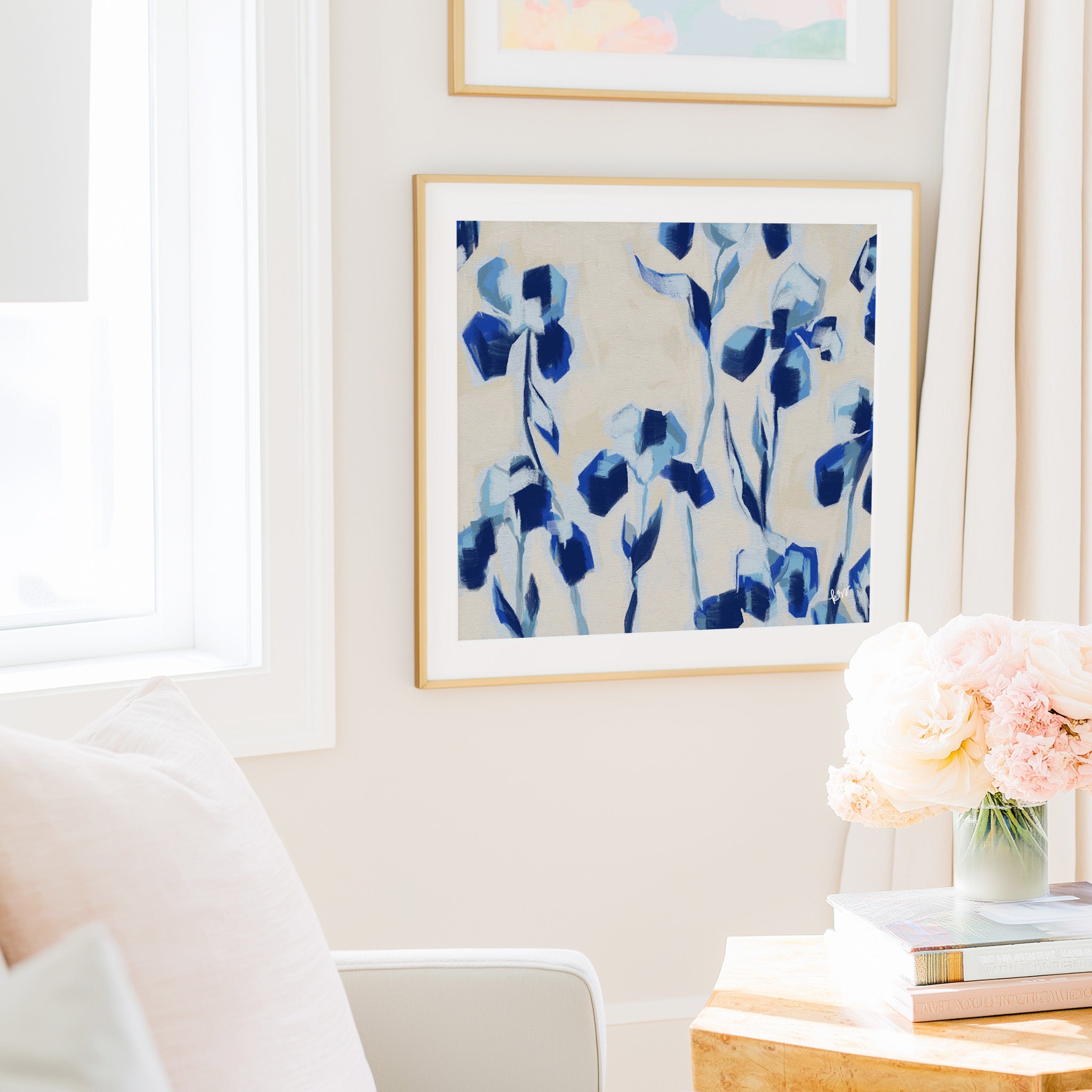 a cozy living room with a white sofa, a framed abstract painting of blue flowers hanging on the wall, and a vase of pink flowers on a wooden table.