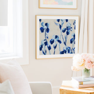 a cozy living room with a white sofa, a framed abstract painting of blue flowers hanging on the wall, and a vase of pink flowers on a wooden table.