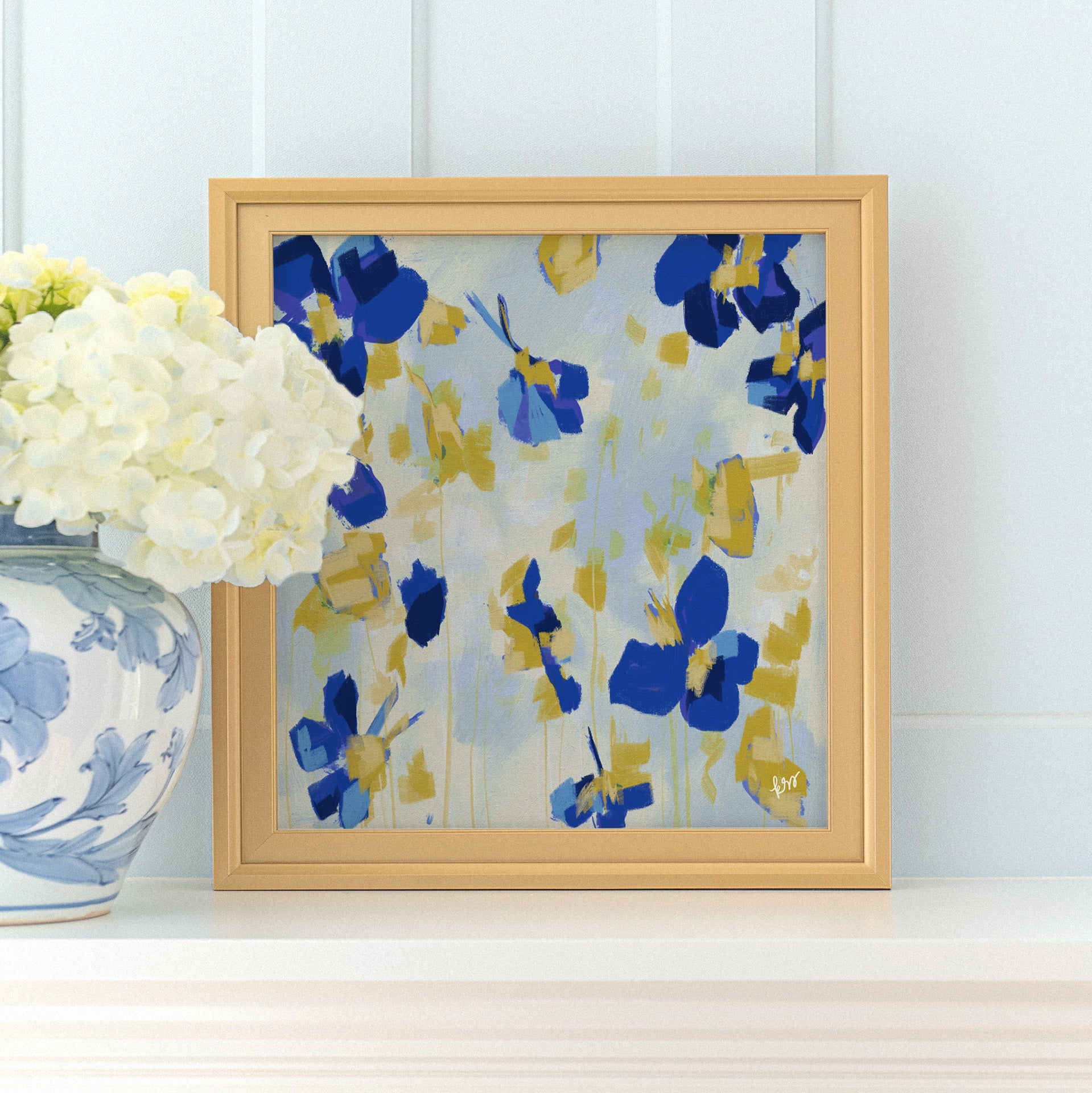 a framed painting of blue and yellow flowers on a white shelf, with a vase of white hydrangeas placed next to it.