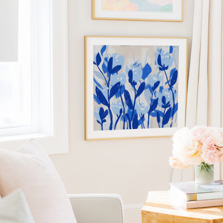 a cozy living room with a white sofa, a framed artwork of blue flowers on the wall, and a vase of pink flowers on a wooden table.