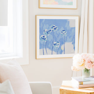 a cozy living room with a framed painting of blue flowers hanging on the wall, a vase of pink flowers on a wooden table, and a stack of books on the table.